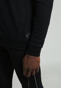 Felpa nera con maniche lunghe, polsi a costine e un piccolo logo sul bordo inferiore. Il tessuto appare morbido con una finitura opaca.