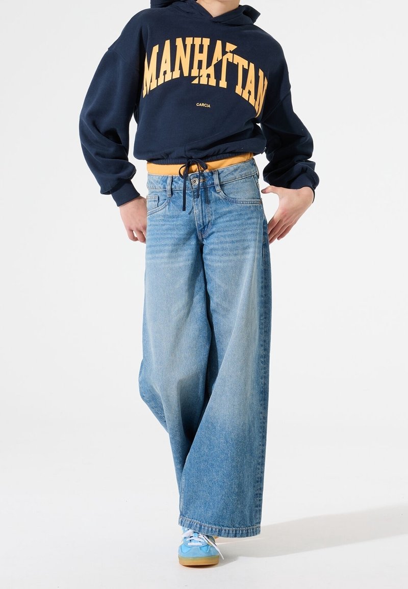 Personne portant un sweat à capuche bleu marine avec le texte « MANHATTAN », un jean large bleu clair et des baskets bleues sur un fond blanc uni.