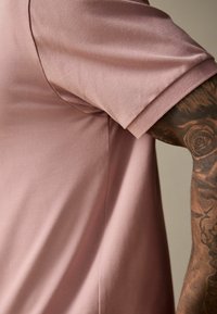 Svetloružová košeľa s krátkym rukávom z textúrovanej látky a rebrovaným golierom, ktorá má bočný šev a ruku s komplikovanými tetovaniami.