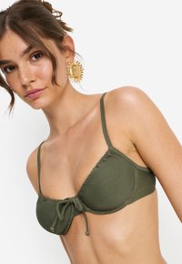Επάνω μέρος μπικίνι σε ελιά πράσινο με ρυτιδωτό σχέδιο και ρυθμιζόμενο κορδόνι μπροστά. Λεπτές τιράντες και λείο ύφασμα με μινιμαλιστικό στυλ.