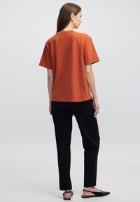 T-shirt arancione bruciato a maniche corte visto da dietro, abbinato a pantaloni neri slim e scarpe nere piatte, texture del tessuto liscia.