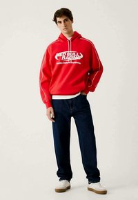 Moletom vermelho com texto branco "RED BULL RACING", com detalhes brancos. Combinado com jeans azul-escuro e ténis brancos. Design simples.