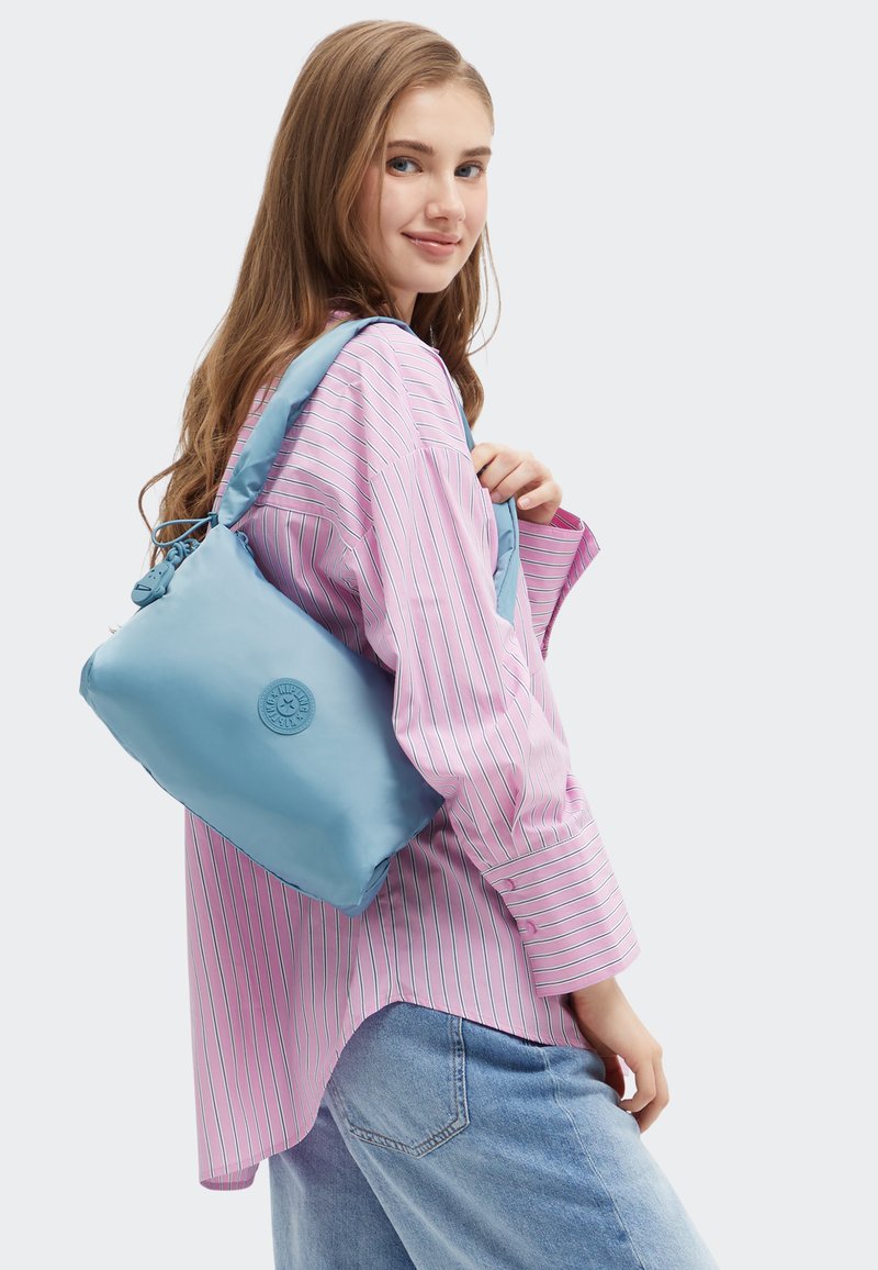 Niebieska nylonowa torba na ramię z teksturowaną naszywką z logo, z regulowanymi paskami i miękkim wykończeniem, noszona na różowej w pasiaki koszuli.