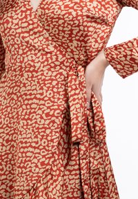 Punainen ja beige kietaisumekko, jossa on teksturoitu eläinkuos, pitkät hihat ja sidottu vyötärö. Sujuva kangas, jossa on virtaava muotoilu.