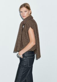 Poncho lavorato a maglia marrone con un design aperto, abbinato a jeans in denim scuro. Presenta un tessuto testurizzato e fessure laterali per facilitare i movimenti.