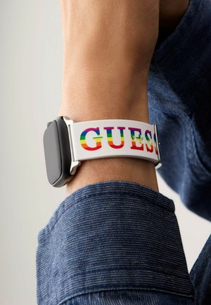 Zegarek na nadgarstek z czarną ramką, białym silikonowym paskiem z napisem "GUESS" w tęczowych literach, noszony na mankiecie niebieskiej koszuli dżinsowej.