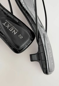 Juodi plokšti batai su krokodilo tekstūra, turintys elegantišką dizainą ir minimalų kulną. Vidpadis pažymėtas "NEXT Forever Comfort."