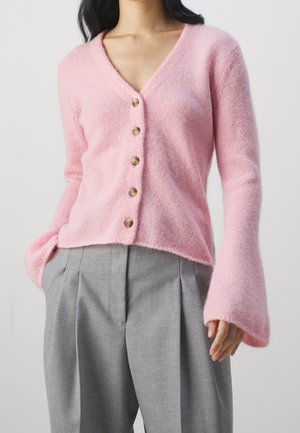 Rosa Strickjacke aus weichem Material, mit V-Ausschnitt, sieben Holzknöpfen und Glockenärmel, kombiniert mit grauen, plissierten Hosen.