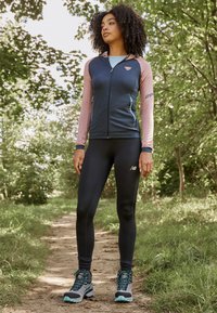Sportliches Outfit mit einer navyblauen und pinken Zip-Jacke, schwarzen Leggings und grauen Wanderschuhen mit türkisfarbenen Akzenten, auf einem üppigen Wanderweg im Freien.