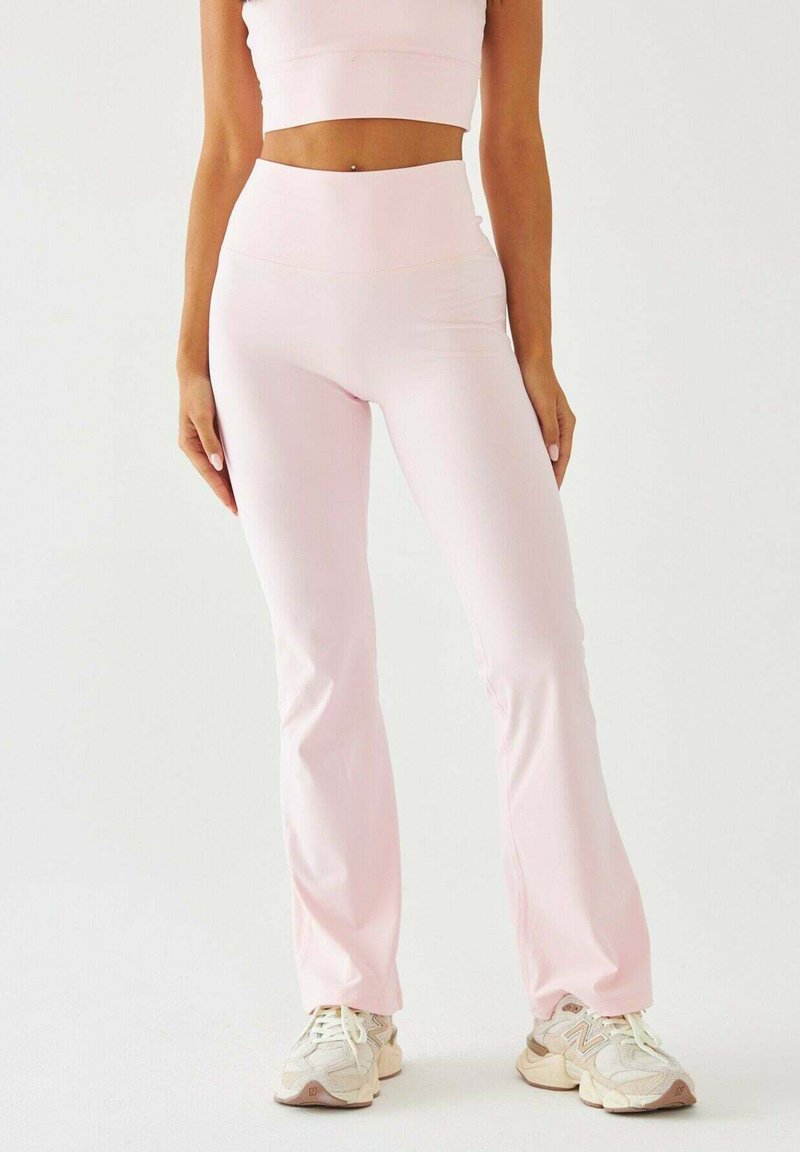 Jasnoróżowy zestaw sportowy składający się z krótkiego topu i spodni z wysokim stanem i rozszerzanymi nogawkami, wykonany z gładkiego, elastycznego materiału. Całość dopełniają casualowe trampki.