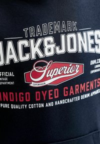 Λογότυπο και κείμενο Jack & Jones σε σκούρο ύφασμα που τονίζουν τα "Superior,", "Indigo Dyed Garments," και "Pure quality cotton and handcrafted denim apparel."