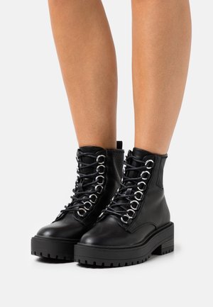 Zwarte leren veterschoenen tot de enkel met dikke zolen, gedragen op blote benen tegen een witte achtergrond.