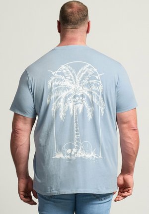 Mann trägt hellblaues T-Shirt mit weißem Motiv einer Palme und Totenköpfen auf dem Rücken, steht vor einfarbigem Hintergrund.
