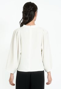 Biela blúzka s nafúknutými rukávmi, vyrobená z hladkej látky. Zadná časť má zips a minimalistický dizajn, skombinovaná s čiernymi nohavicami.