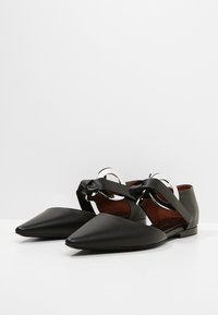 Bailarinas de cuero negro con punta afilada, que presentan un diseño de tira y herrajes circulares plateados. Textura suave y silueta de perfil bajo.