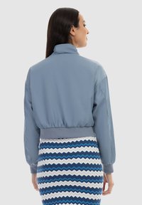 Jachetă bomber de culoare albastru deschis, cu un brâu și manșete gri cu model texturat. Modelul poartă o fustă cu un model zigzag în albastru și alb, văzută din spate.