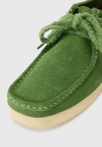 Žalia suede mokasinų avalynė suapvalintu priekiu, tekstūruotomis raištelėmis ir kontrastiniu smėlio spalvos padu. Išsami siuvinėta apdaila palei kraštą.