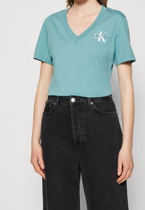 Kobieta w turkusowej koszulce w serek od Calvin Klein Jeans, włożonej w czarne dżinsy z wysokim stanem, stojąca na jednolitym tle.