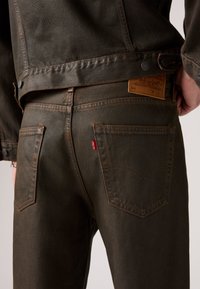 Temno zelena jeans jakna in kavbojke s bakrenim šivom, vidnimi zadnjimi žepi in usnjenim logotipom na pasu.