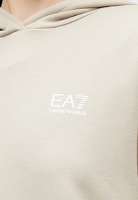 Gaiši bēša sporta jaka ar mīkstu tekstūru, uz krūtīm redzams balts EA7 Emporio Armani logotips, ar vaļēju kapuces dizainu.