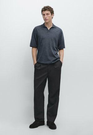 Young man standing with hands in pockets wearing dark gray short-sleeve collared shirt and black wide-leg trousers on plain background.