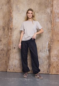 Blondynka stojąca oparta o drewnianą ścianę, ubrana w beżowy T-shirt, luźne czarne spodnie i brązowe trampki z białymi paskami.