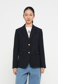 Marineblazer met twee zijzakken en gouden knopen, voorzien van een getailleerde pasvorm en reverskraag, gecombineerd met lichtblauwe jeans.