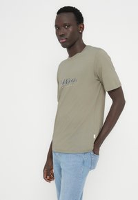 Λαδί πράσινο μπλουζάκι με στρογγυλή λαιμόκοψη, κοντά μανίκια και γραφικό σχέδιο μπροστά. Συνδυάζεται με τζιν ανοιχτού μπλε χρώματος, το ύφασμα έχει λεία υφή.