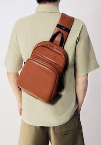 Sac en cuir marron en bandoulière avec des compartiments zippés, une sangle ajustable et une texture lisse ; porté dans le dos d'une personne en tenue décontractée.