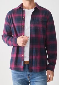 Camisa de franela en patrón de cuadros en tonos azul marino y burdeos, con diseño abotonado, bolsillo en el pecho y acentos de botones en color dorado.