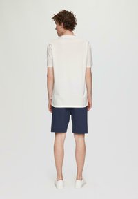 Camiseta blanca de manga corta combinada con pantalones cortos azul marino, vista desde atrás. El modelo lleva zapatillas blancas. Tela suave y diseño informal.