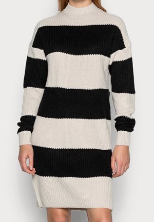 Kvinde iført en sort og hvid strikket kjole med brede, vandrette striber, lange ærmer og høj halsudskæring.