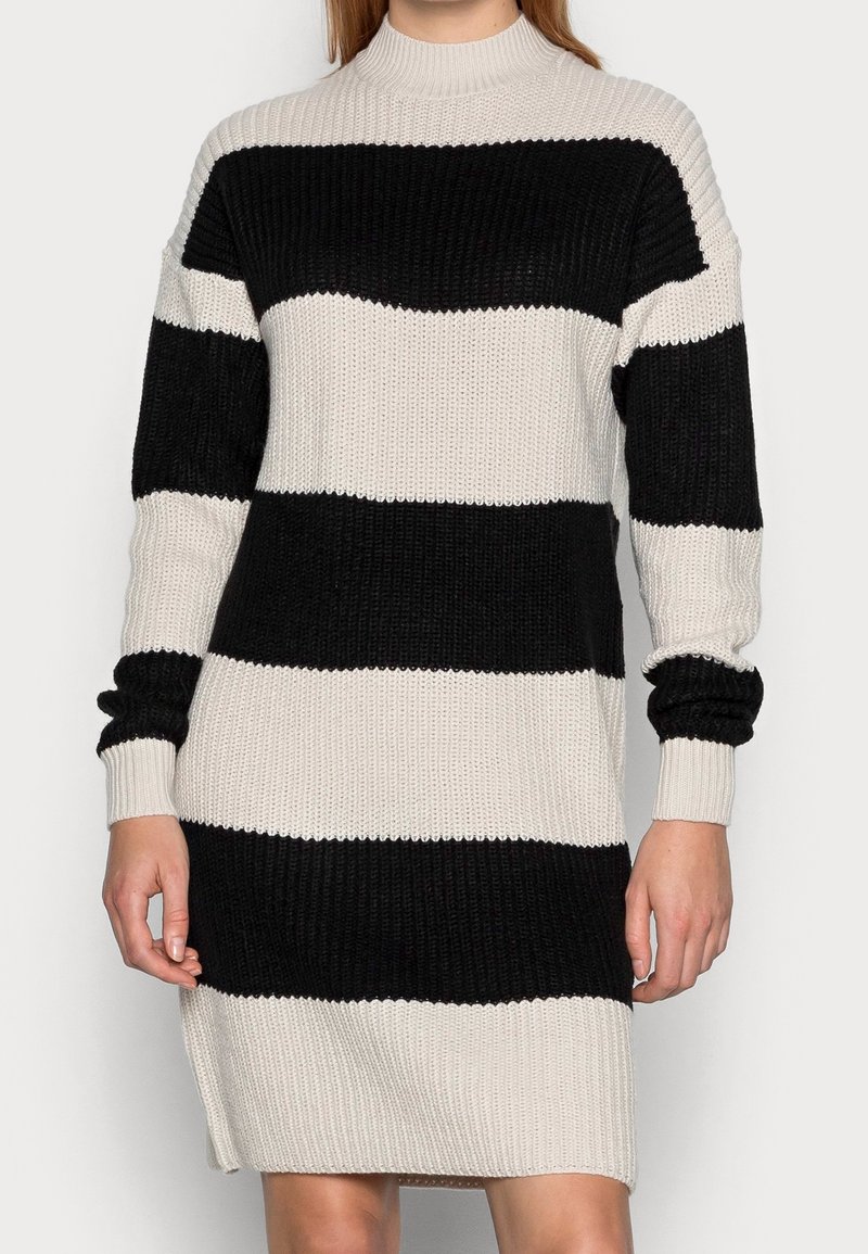 Femme portant une robe pull tricotée à larges rayures horizontales noires et blanches, manches longues et col haut.