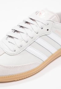 Zapatilla deportiva blanca de cuero con tres rayas blancas, puntera de ante gris texturizado, cordones gris claro y una suela de goma. Presenta la marca "Samba".
