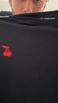 Črna majica s kratkimi rokavi iz gladkega materiala, na sprednji strani ima majhen rdeč motiv višnje. Preprost dizajn.