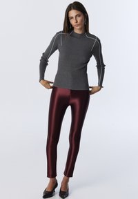 Woman with straight brown hair wearing gray ribbed long-sleeve top with white seams, burgundy shiny leggings, black heels, standing against white background.