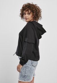 Sudadera negra corta con capucha ajustable, con mangas anchas y puños de canalé, combinada con pantalones cortos de denim claro.