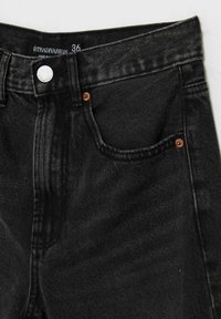 Primer plano de unos jeans de mezclilla negros, mostrando el botón, los lazos del cinturón, el bolsillo frontal con remaches de cobre y la etiqueta indicando talla 36.