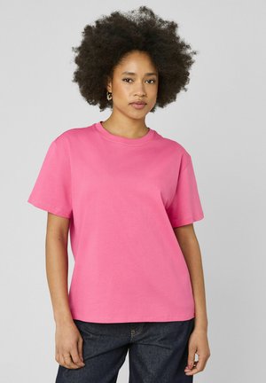 Jeune femme aux cheveux afro naturels portant un t-shirt rose vif à manches courtes et un jean foncé, debout devant un fond uni.