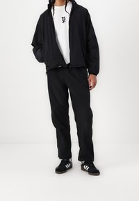 Chaqueta ligera negra con puños elásticos, combinada con una camiseta blanca con un estampado gráfico y pantalones negros de corte holgado. Zapatillas negras.