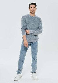 Pull gris à côtes avec un col rond, associé à un jean bleu clair et des baskets blanches. Le modèle se tient de manière décontractée contre un fond uni.