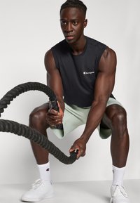 Camiseta atlética sin mangas negra, pantalones cortos verdes claros, zapatillas blancas, agarrando una cuerda de batalla negra, expresión concentrada, físico tonificado, fondo minimalista.