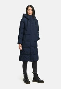Námornícky puffer kabát s kapucňou, ktorý má dlhý strih, veľké vrecká, zips a textúrovaný prešívaný vzor. Nosený s čiernymi čižmami.
