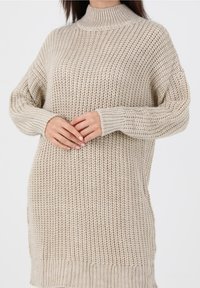 Pull en maille beige avec un col montant, des poignets côtelés et une coupe ample. La texture est grossière avec un motif de points ouverts.