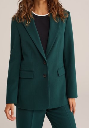Vrouw draagt een op maat gemaakte donkergroene blazer met twee knopen en bijpassende broek, gecombineerd met een zwart-wit rondhals shirt.