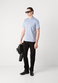Lys blå t-skjorte med korte ermer, svarte slim-fit bukser, svart skinnjakke over armen, og svarte dressko. Rektangulære solbriller på.