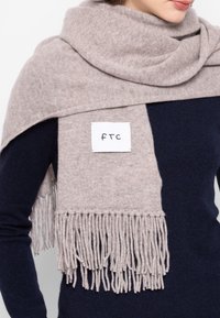 Jasnoszary wełniany szalik z frędzlami na krawędziach, zarzucony na granatowy sweter. Przyszyta w środku metka z napisem „ftc”.