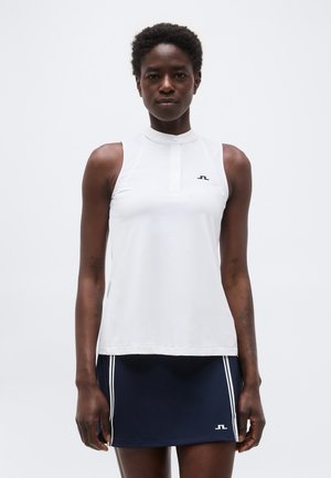 Άτομο που φοράει άσπρο τοπ χωρίς μανίκια με ψηλό γιακά και μπλε φούστα με άσπρη διακόσμηση, που στέκεται μπροστά σε απλό φωτεινό φόντο.