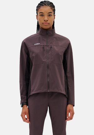 Donna con capelli intrecciati che indossa una giacca viola scuro con zip e pantaloni abbinati, in piedi davanti a uno sfondo bianco uniforme.