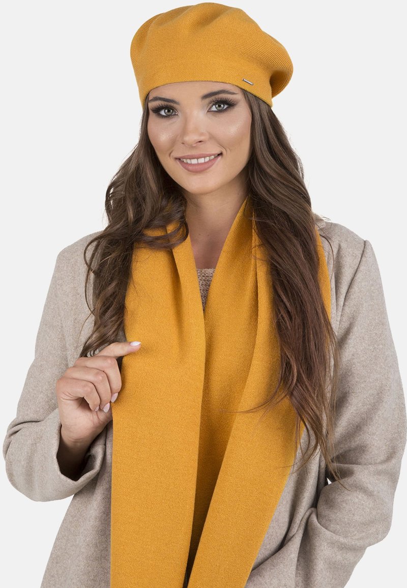 Boina de punto amarilla y bufanda a juego llevadas sobre un abrigo beige; textura suave, diseño sencillo y sin patrones ni elementos metálicos visibles.